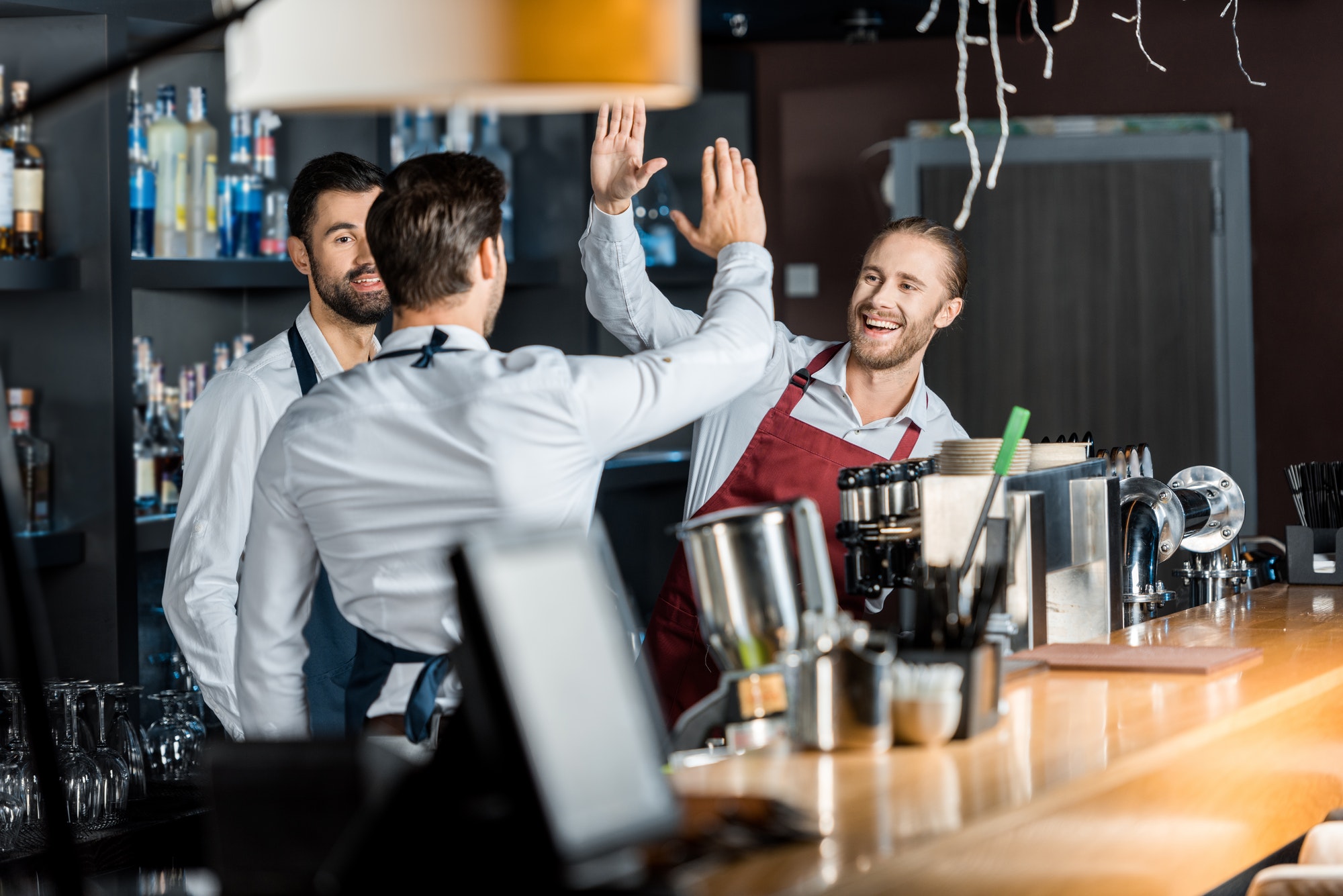 handsome smiling barmen in aprons high five at workplace